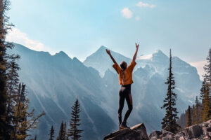 Woman is standing in front of mountains and raising her arms